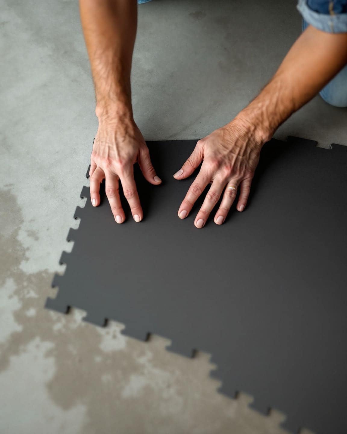Hands lay black interlocking rubber tiles on a concrete floor.