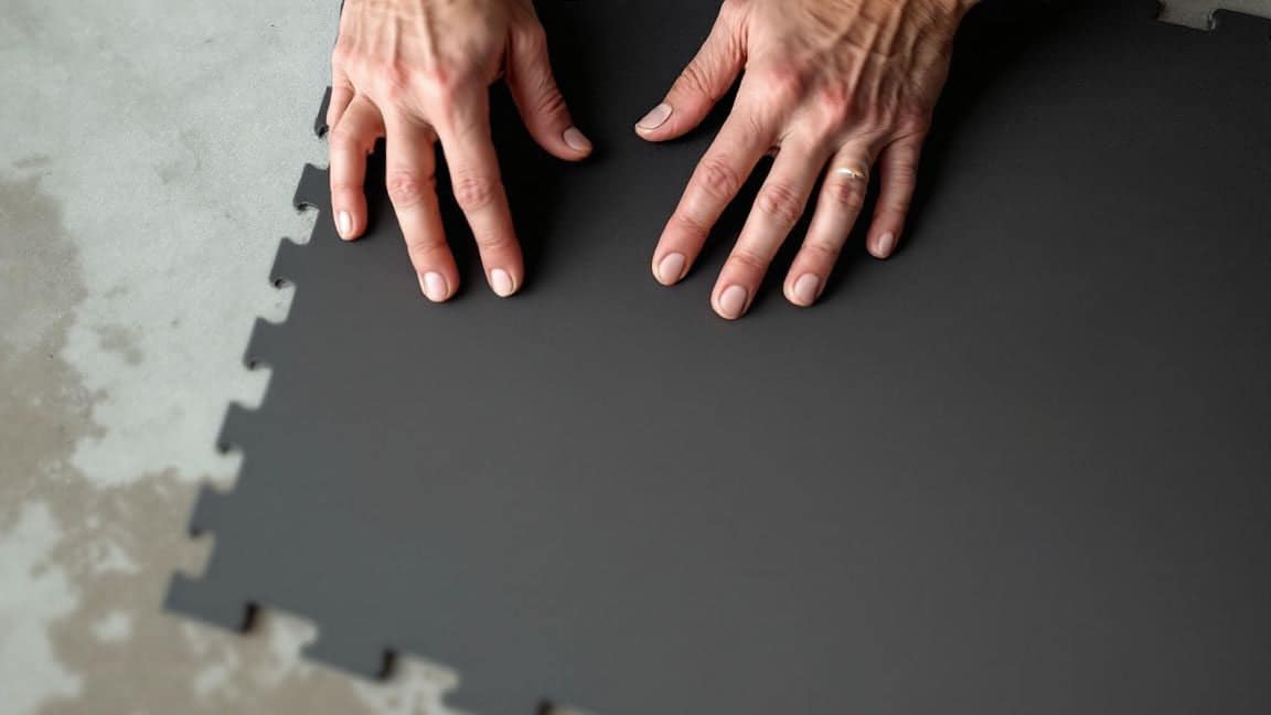 Hands lay black interlocking rubber tiles on a concrete floor.